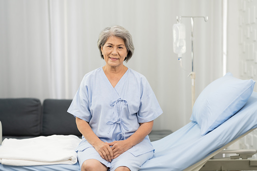 Senior asian woman sitting on bed with receiving saline solution from bag at hospital ward. medicine, health care, old people and quarantine concept