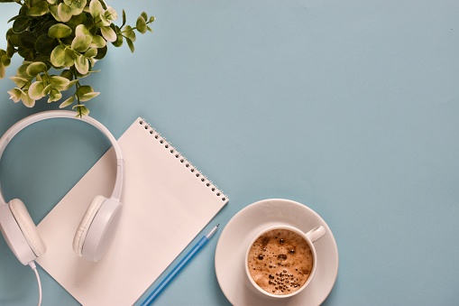 Headphones with blank notepad, cup coffee, green plants and pen on a light blue background. Workspace. Flat lay composition with copy space