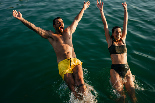 Spontaneous and fun young adult couple jumping in the sea, enjoying their vacation, smiling in their bathing suit. Screaming and laughing loud. Captured vacation moment, to remember