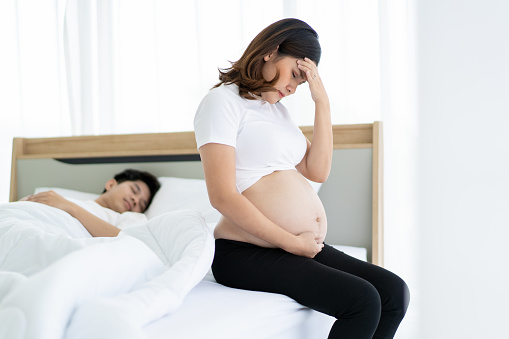 Upset and crying pregnant woman sitting on the bed while her husband sleeping. Depressing suffer in pregnant woman with her husband. Problem in marriage life between husband and wife.