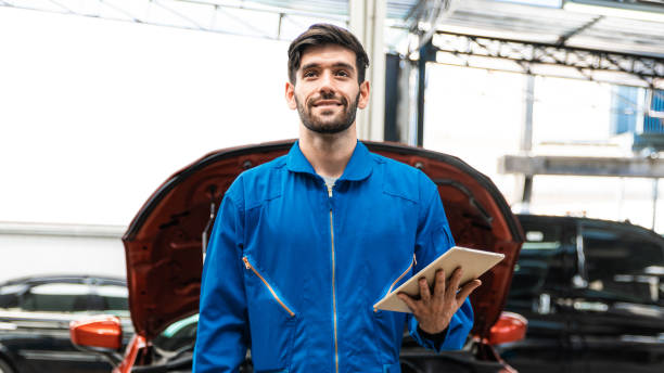 mecânico em uniforme azul workwear segurando tablet e olhando para cima com carro desfocado ao fundo. serviço de reparação de automóveis, trabalho em equipe de ocupação profissional. manutenção do veículo. - repairing computer work tool conformity - fotografias e filmes do acervo