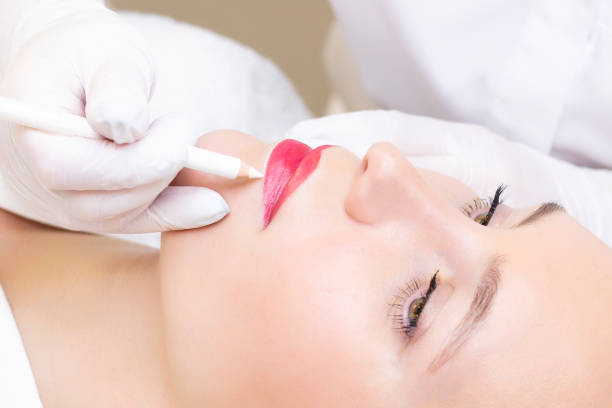 istock Young woman having permanent makeup on her lips at the beauticians salon. Permanent Makeup (Tattoo). drawing a contour with a white lip pencil 1408587604