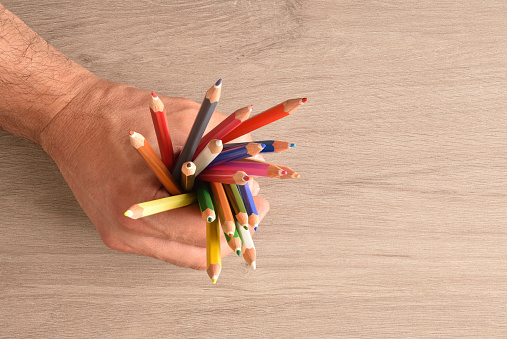 Hand of a man holding a group of colored pencils on a wooden table. Top view