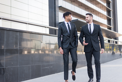 two businessmen wearing suits walking and smiling and talking outdoor in a business area.