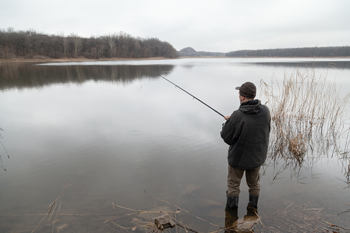 A fisherman in boots stands in the water of a calm river and holds a fishing rod. In front of him is the expanse of a wide river. Trees grow along the banks. View from the back