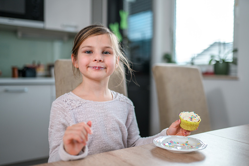 Waist up shot of smiling mischievous little girl. She is smiling while eating a cupcake at the kitchen table.