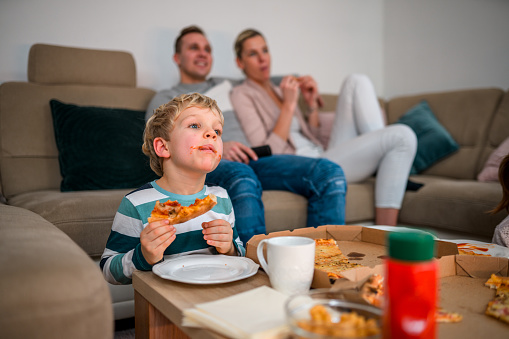 Front shot of little blond hair boy with sauce in his mouth eating pizza. He is at the living room watching a movie with his parents.