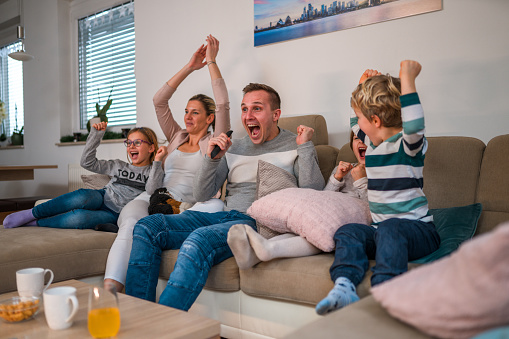 Cheerful white family seated in living room. Mid adult couple and kids celebrate match game score by lifting arms in the air.