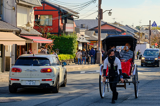 Kyoto, Japan - November 19, 2021: A rickshaw driver carries two passengers along the main thoroughfare in Arashiyama. Rickshaw rides are among the activities allowing tourists to experience traditional Japanese culture in Kyoto, along with tea ceremonies, kimono rentals, and maiko performances.