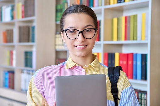 Portrait of high school student looking at camera in library. Smiling teenage girl in glasses with backpack and laptop in her hands. Study, education, knowledge, adolescence concept