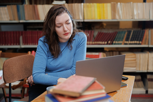 Female adult student using laptop for for e-learning at the desk with a stack of selected books in the library