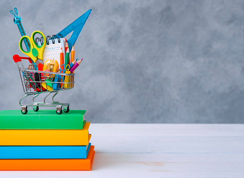 Colorful school supplies in the shopping basket on a gray background with a copy of the text space. A stack of books with colorful covers. The concept of returning to school for the new academic year.