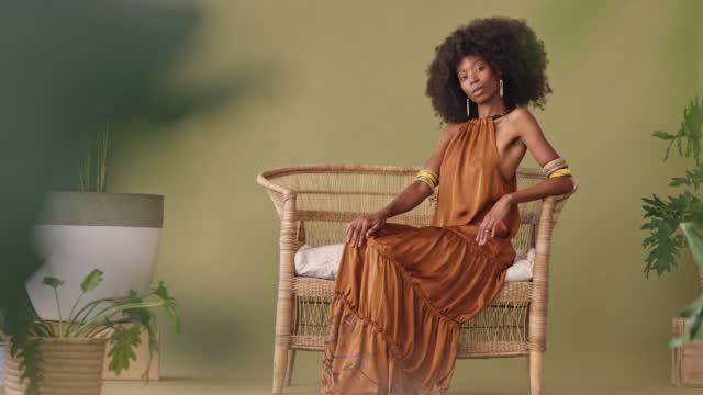 Full length beautiful African American woman with an afro sitting alone on a wicker chair and feeling confident. Powerful and exotic black woman with an afro feeling like a queen while posing in the studio