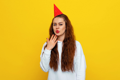 Oops. Embarrassed young woman in a celebratory cap, grimacing with awkward expression, being guilty and hoping that nobody will know about her fault. Colorful studio portrait with yellow background