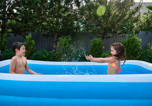 Happy cheerful caucasian children splashing each other with water while playing in an inflatable swimming pool in the yard of private house, enjoying summer holidays on a beautiful warm and sunny day