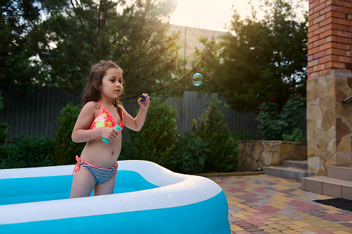 Happy funny child, adorable little girl in a bathing suit blows soap bubbles while sunbathing in an inflatable pool in the backyard garden of the house, at sunset. Summer vacation and leisure concept