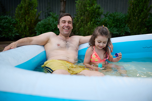 Happy young Caucasian man, dad and his adorable little girl, having water fun together, sitting in the inflatable swimming pool at home backyard garden and enjoying time together during a weekend