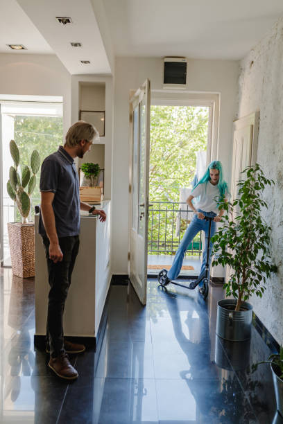 Young man standing in the hall, his female flatmate coming home on push scooter Young man standing in the hall of the apartment, his female flatmate entering home on e-scooter after work, sustainable commuting flatmate stock pictures, royalty-free photos & images