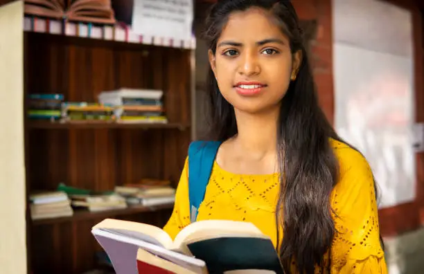 Photo of Indian female university student reading a book near outdoor library at campus.