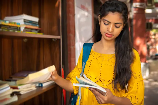 Photo of Indian female university student reading a book near outdoor library at campus.