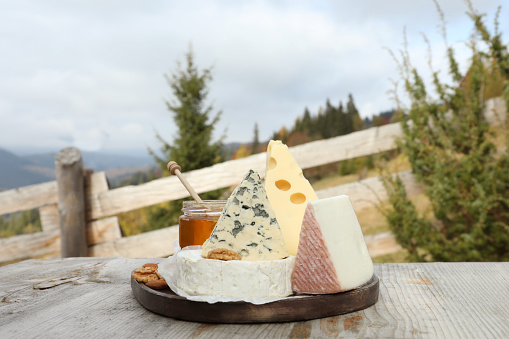 Different types of delicious cheeses on wooden table against mountain landscape