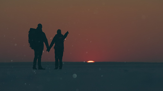 The two travelers standing through the snow field against beautiful sunset