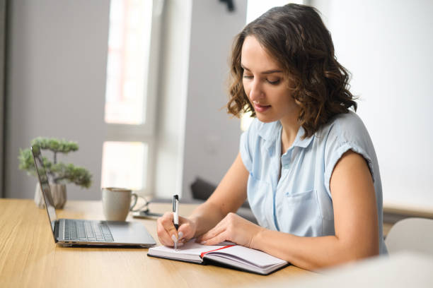 istock Caucasian young woman is using laptop computer for remote work or studying online, she takes notes watching webinars or classes at kitchen table 1408642746
