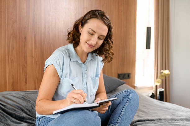 istock Charming caucasian woman sitting at the bed at home, taking notes, learning. Smiling business lady analyzing tasks while works in the home 1408642811