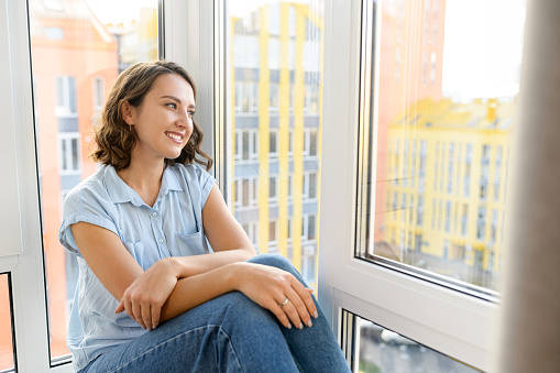 Dreamy full of energy woman in casual clothes resting near the window, sitting on the windowsill in modern living room. Lady posing and looking away
