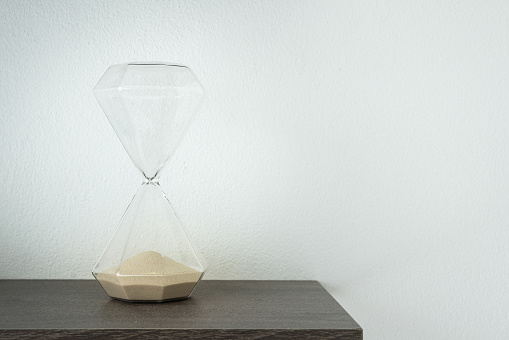 A sandglass or hourglass is placed on wooden shelf. Interior decoration object photo, selective focus.