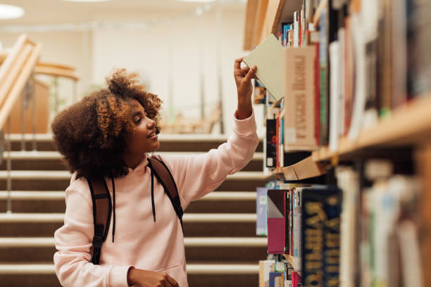 chica tomando un libro de la estantería en la biblioteca - biblioteca fotografías e imágenes de stock