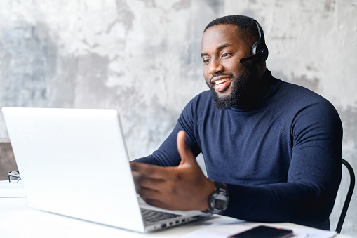 Friendly African-American male entrepreneur wearing wireless headset using a laptop for online communication with customers, a dark skinned employee using hands free device for video conversation