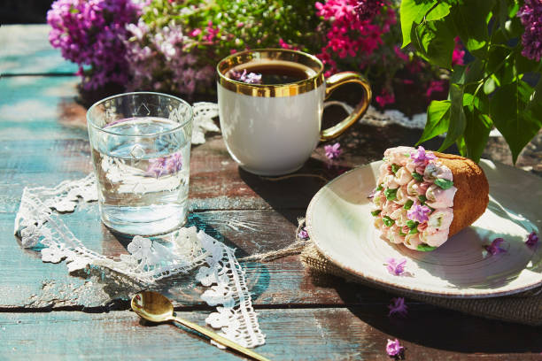 branche extérieure d’été avec une tasse de café, un beau cupcake, un verre d’eau, des fleurs et une décoration en dentelle. pique-de base de chalet avec des tons d’été lumineux de fleurs par une journée ensoleillée. ombres dures - snack coffee instant coffee cappuccino photos et images de collection