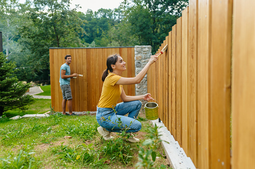 Photo of a young couple painting a picket fence and little wooden house in a yard.