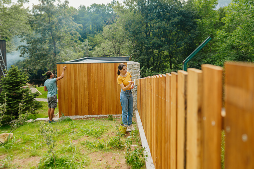Photo of a young couple painting a picket fence and little wooden house in a yard.