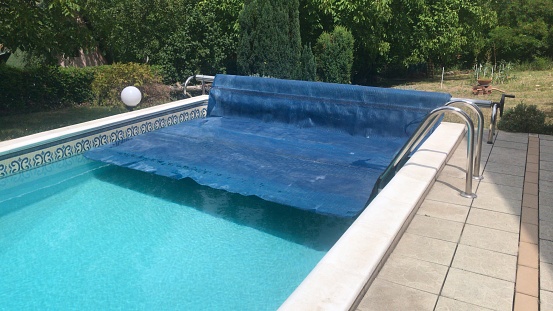 Angled perspective view of half removed blue solar pool cover with clean swimming pool water and stainless steel ladder