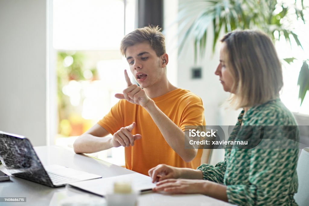Deaf teenage boy signing while e-learning from home Waist-up view of high school student’s online education with parent assistance. Part of lifestyle series depicting deaf family and friends. Deafness Stock Photo