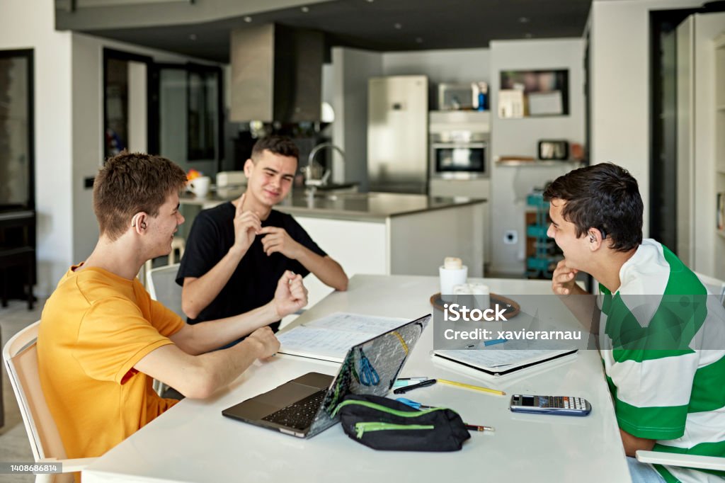 Boys sitting at dining table doing homework and signing Deaf teenagers in casual attire taking a break from their assignments to communicate and laugh. Part of lifestyle series depicting deaf family and friends. Deafness Stock Photo