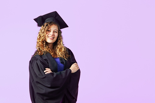 Happy, smiling young woman with long curly hair, wearing academic attire, beams at the camera, with plenty of copy space behind her.