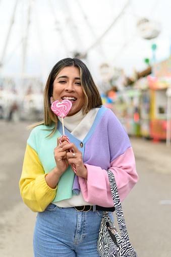 young girl in casual clothes eating a delicious lollipop while in an amusement park looking at a smiling camera