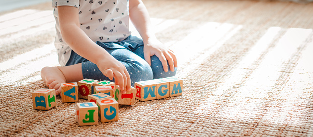 3 year old child plays with wooden cubes with colorful letters on the floor in the room a little girl is building a tower at home or in the kindergarten. Educational toys for young children.