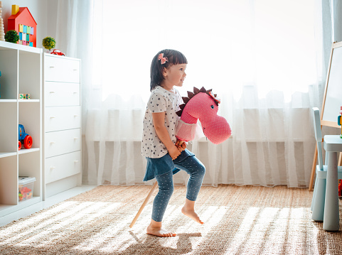 little girl jumps and runs on a horse stick in the nursery. Handmade. Child 3 years of fun playing active games in kindergarten. Physical child development