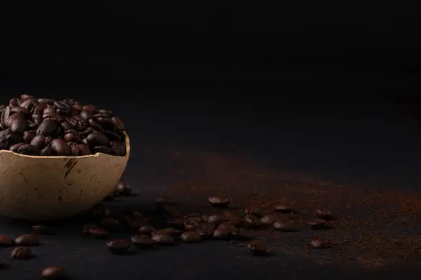 A bowl is full of coffee beans on a black surface with scattered coffee beans leaving space to place text or design. Flat front view