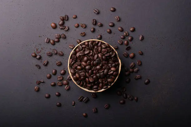 Bowl full of coffee beans on a black surface with scattered coffee beans. Aerial view plan.