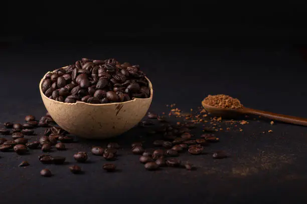 A bowl is full of coffee beans accompanied by a wooden spoon with freeze-dried coffee on a black surface with scattered coffee beans. Flat front view