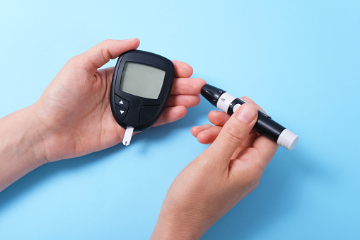 Woman using glucometer, checking blood sugar level. Diabetes concept on blue background. The hand holds the glucometer and pricks the finger with a lancet