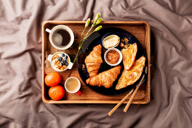 Continental breakfast on wooden tray in bed - coffee and croissants - from above (top view). Continental breakfast on rustic wooden tray in bed (grey sheets). Coffee, croissants, jam, butter, fruits and flowers. Lazy, romantic, weekend morning meal in a hotel suite or home. bed and breakfast stock pictures, royalty-free photos & images