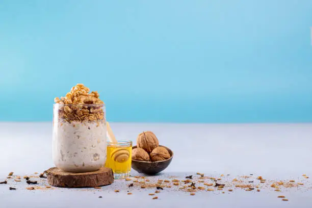 Overnight oats with walnuts, accompanied with honey and whole walnuts. Photo with a blue background and space to add text. Cinnamon powder, oatmeal, and cloves are scattered on the surface.