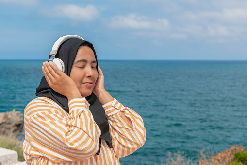 Islamic woman wearing a hijab and listening to music on her headphones on an out of focus sea background. Selective focus. Music concept.