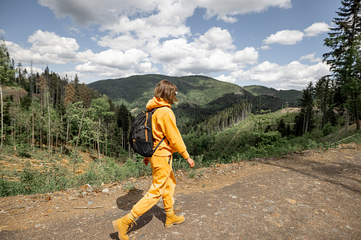 Young person in orange sports suit and backpack walks on the mountain path in pine forest, side view. Concept of traveling in the mountains